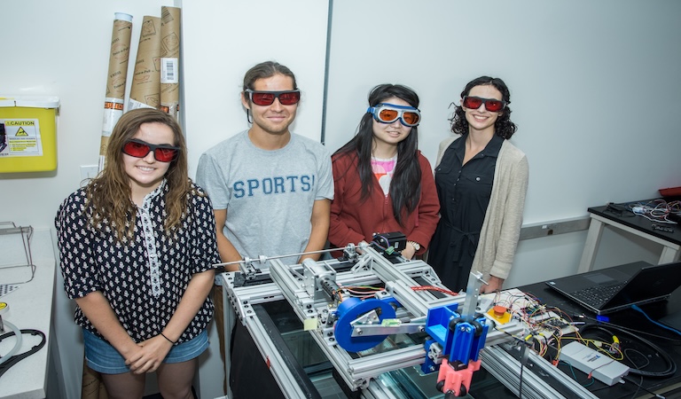 One professor and four students stand close together wearing protective eyeglasses and smiling at the camera. In front of them there are components of a mechanism they built.
