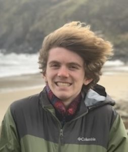 Student Jimmy Boyle on windy beach with surf in background and wearing a multi-green Columbia brand jacket